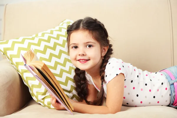 Menina bonita sentada no sofá com livro, em casa fundo interior — Fotografia de Stock