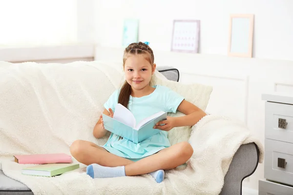 Hermosa niña sentada en el sofá con libro, en el fondo interior de casa —  Fotos de Stock