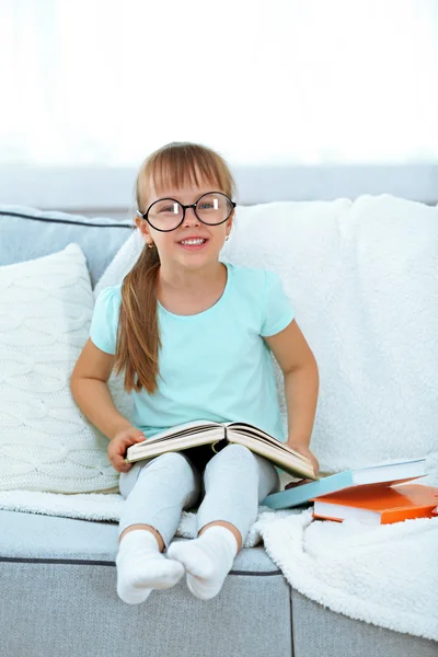 Hermosa niña sentada en el sofá con libro, en el fondo interior de casa — Foto de Stock