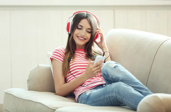 Mujer escuchando música en auriculares mientras está acostada en el sofá en la habitación —  Fotos de Stock