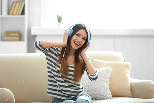 Mulher ouvindo música em fones de ouvido enquanto se senta no sofá no quarto — Fotografia de Stock