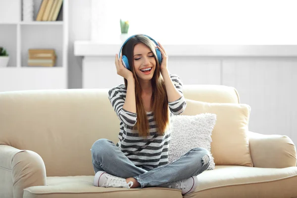 Woman listening music in headphones while sitting on sofa in room — Stock Photo, Image