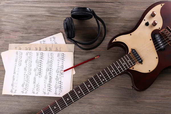 Electric guitar with headphones and music notes on wooden table close up — Stock Photo, Image
