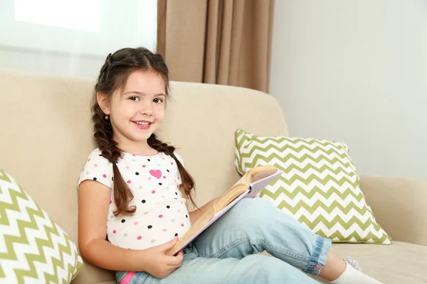 Frumoasă fetiță așezată pe canapea cu carte, pe fundal interior acasă — Fotografie, imagine de stoc