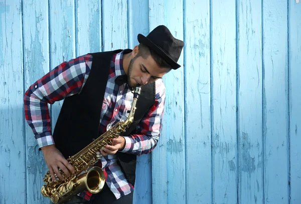 Hombre guapo en sombrero toca saxo sobre fondo de madera azul —  Fotos de Stock