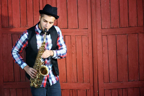 Handsome happy man in hat plays music on sax in front of red wooden background — Stock Photo, Image