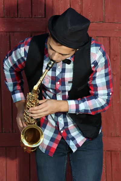 Beau homme heureux dans le chapeau joue de la musique sur le saxophone devant le fond en bois rouge — Photo