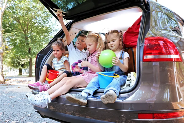 Tres hermosas chicas y un niño se sientan en el maletero de un coche y se ríen —  Fotos de Stock