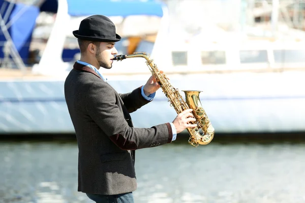Mladý muž hraje na saxofon na břehu řeky — Stock fotografie