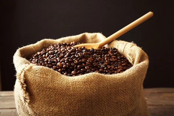 Sac avec grains de café torréfiés avec cuillère sur table en bois devant fond sombre — Photo