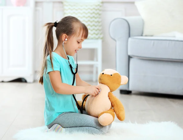 Niña linda con estetoscopio y oso de peluche sentado en la alfombra, en el fondo interior del hogar —  Fotos de Stock