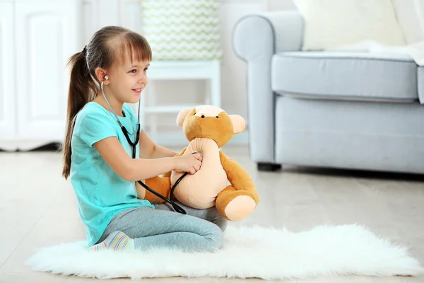 Kleines nettes Mädchen mit Stethoskop und Teddybär auf Teppich sitzend, auf häuslichem Hintergrund — Stockfoto