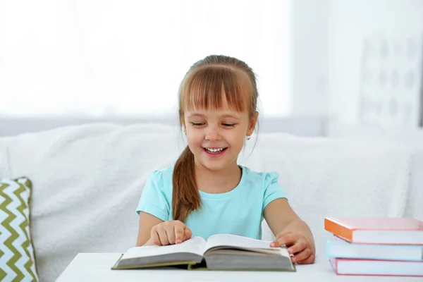 Hermosa niña sentada en el sofá con libro, en el fondo interior de casa —  Fotos de Stock