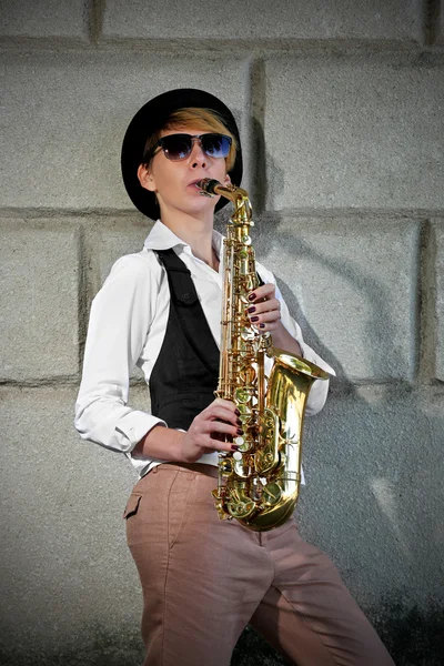 Atrakcyjna kobieta w kapeluszu gra saksofon na tle ściany Cegła — Zdjęcie stockowe