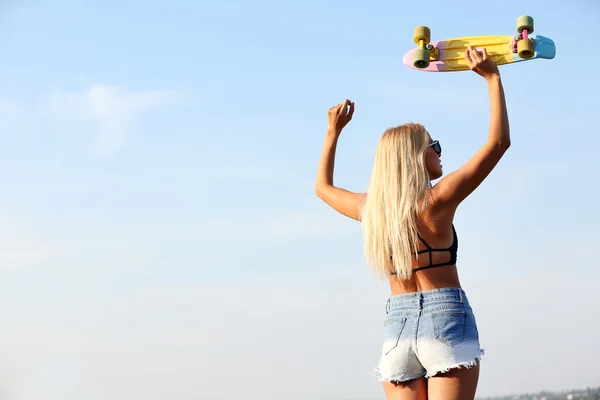 Mujer joven con tabla de patinar sobre fondo azul cielo —  Fotos de Stock