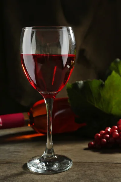 Sklenice na červené víno proti proutěný koš s hroznové víno a láhev na dřevěný stůl — Stock fotografie