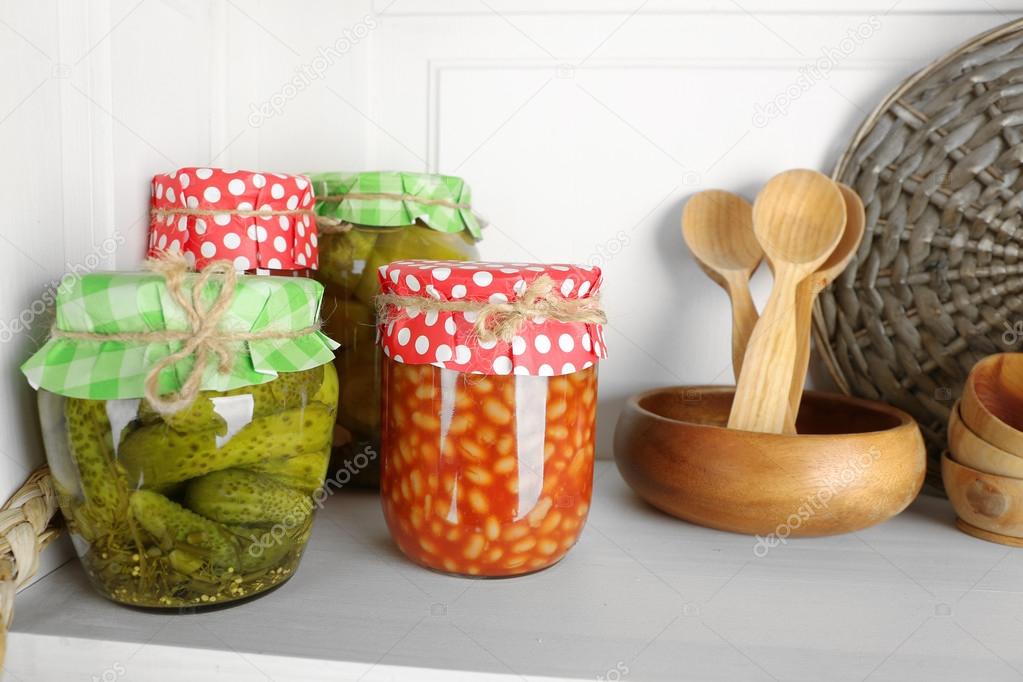 Jars with pickled vegetables and beans, spices, book of recipes and kitchen utensils on shelf