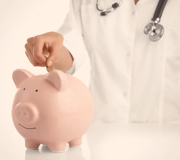 Doctor putting coin into piggy bank — Stock Photo, Image