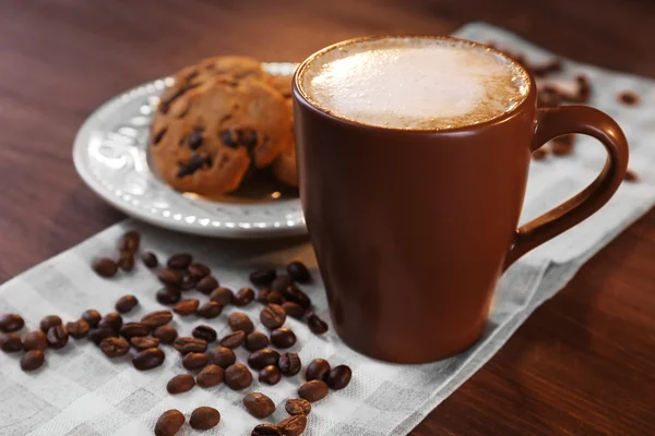 Xícara de café com grãos de café torrados ao redor e biscoito em guardanapo de algodão no fundo de madeira, close-up — Fotografia de Stock