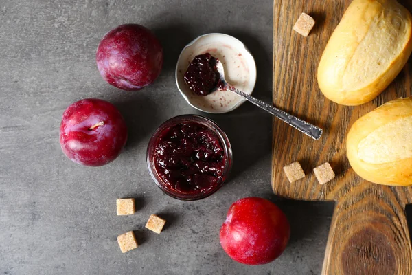 Un barattolo di marmellata saporita, un cucchiaio, prugne, cracker su fondo grigio e focacce fresche su tavoletta di legno — Foto Stock