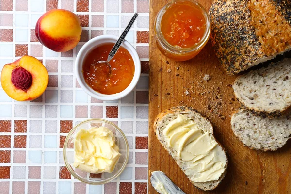 Gustosa marmellata in vasetto e ciotola, pesche mature, pane con burro e tavoletta di legno su fondo mosaico — Foto Stock