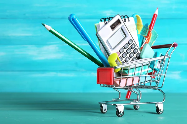 Bright stationery objects in mini supermarket cart on blue wooden background — Stock Photo, Image