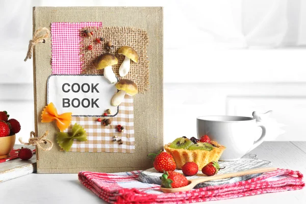 Libro de cocina decorado con taza de té y sabroso pastel sobre fondo blanco —  Fotos de Stock