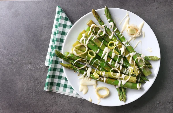 Hidangan asparagus segar dengan irisan bawang di piring putih dengan serbet katun kotak-kotak di meja abu-abu — Stok Foto
