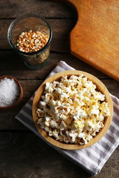 Salted popcorn in a bowl on stripped napkin with board and glass of corns on wooden table — Stock Photo, Image