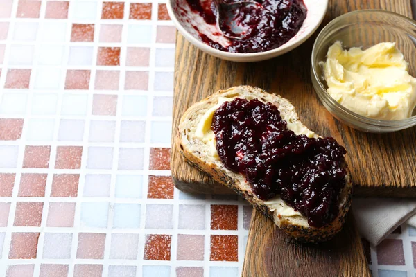 Confettura gustosa sul piatto, burro in una ciotola, pane su tavoletta di legno e un tovagliolo su sfondo mosaico — Foto Stock