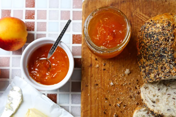 Selai lezat dalam guci dan mangkuk, buah persik matang, mentega, roti segar dan tablet kayu pada latar belakang mosaik close-up — Stok Foto