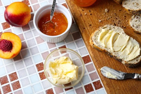Selai lezat dalam guci dan mangkuk, buah persik matang, roti dengan mentega dan tablet kayu dengan latar belakang mosaik — Stok Foto