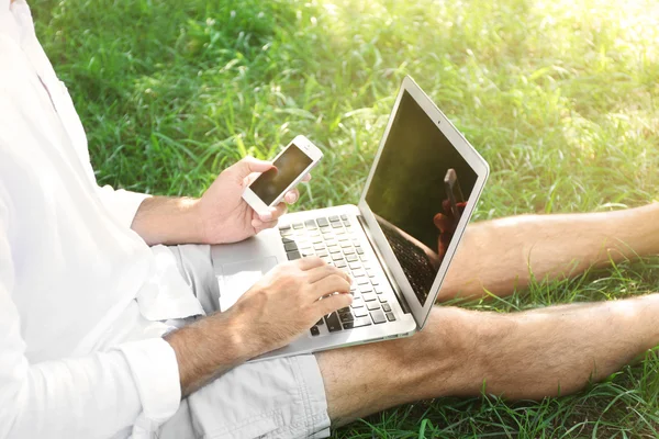 Hombre con portátil y teléfono celular sentado en la hierba verde en el parque, de cerca — Foto de Stock