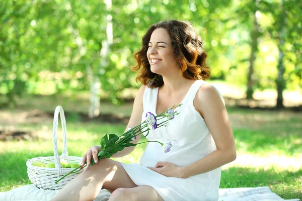 Atraktivní těhotná žena s květinami v rukou na bílou deku v parku — Stock fotografie