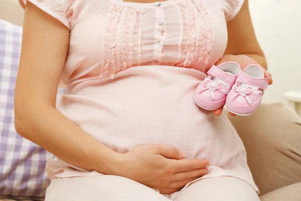 Mulher grávida feliz bonito no sofá aconchegante com botas de bebê no quarto, close-up — Fotografia de Stock