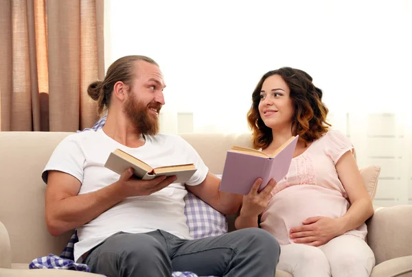 Ευτυχισμένο ζευγάρι αγαπημένη ανάγνωση βιβλίων στην αναμονή για την γέννηση του μωρού στον καναπέ στο δωμάτιο — Φωτογραφία Αρχείου