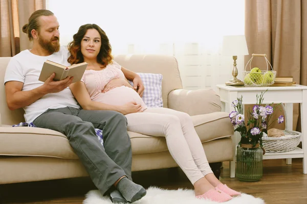 ハンサムな男は、部屋のソファーに彼の素敵な妊娠中の女性に本を読む — ストック写真