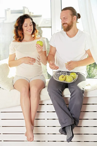 Happy beloved couple in waiting for baby's birth on window board with apples — Stock Photo, Image