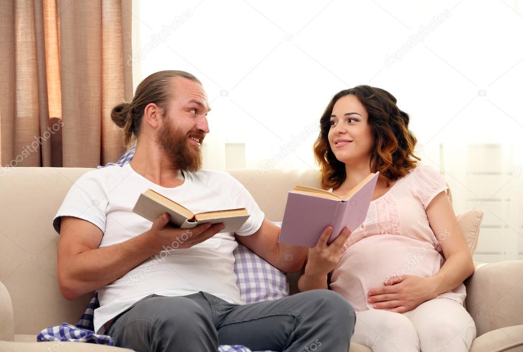 Happy beloved couple reading books in waiting for baby's birth on sofa in the room