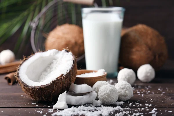 Candies in coconut flakes, glass of milk and fresh coconut on dark wooden background