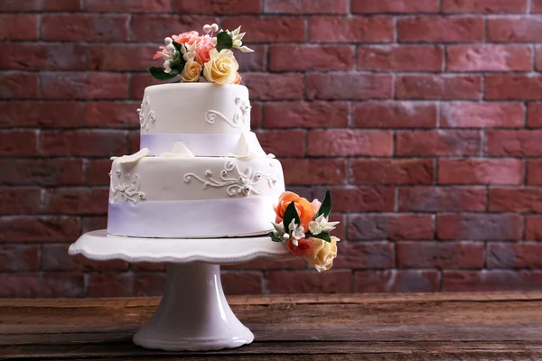 Beautiful wedding cake decorated with flowers on wooden table against brick wall background — Stock Photo, Image