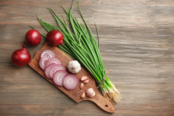 Red onions circles, green onion with garlic on board against wooden background — Stock Photo, Image