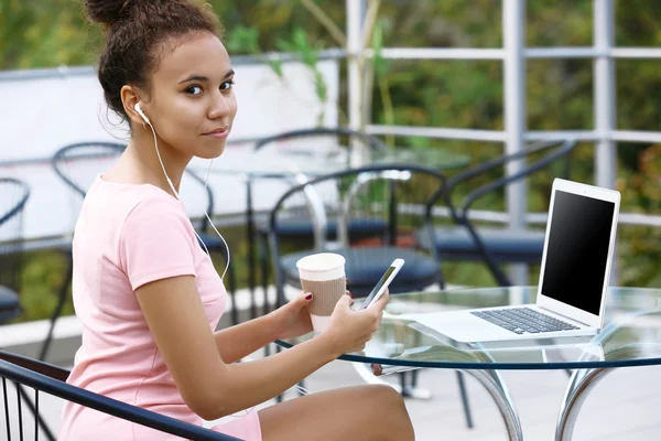 Unga vackra kvinnan i rosa klänning med laptop dricka kaffe och lyssnar musik genom hörlurar på sommarterrassen — Stockfoto