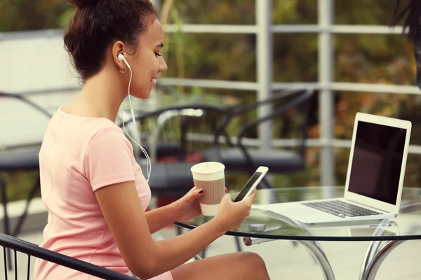 Young pretty woman in pink dress with laptop drinking coffee and listening music by headphones at summer terrace — Stock Photo, Image