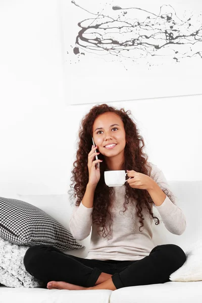 Giovane donna seduta su comodo divano con tazza di caffè in mano e parlando al cellulare — Foto Stock