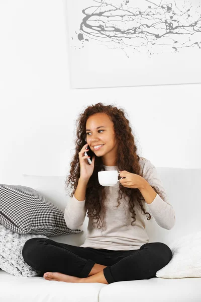 Mujer joven sentada en cómodo sofá con taza de café en las manos y hablando por teléfono celular — Foto de Stock