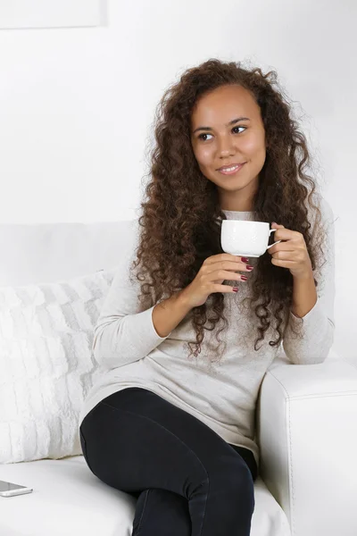 Giovane donna seduta su comodo divano con tazza di caffè in mano in camera bianca — Foto Stock