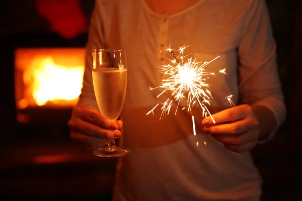 Female hands with sparkler and glass of sparkle wine on fireplace background — Stock Photo, Image