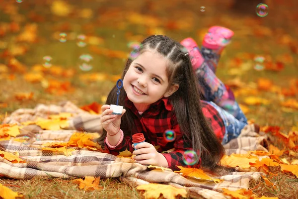 Cute girl lying on plaid and blowing soap bubbles in park — Stock Photo, Image