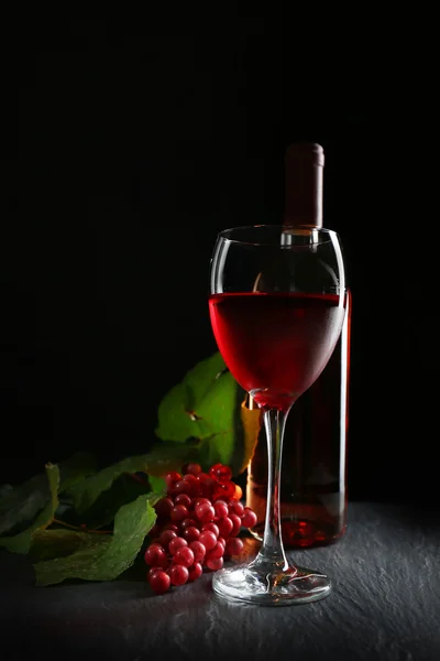 Uma garrafa e um copo de vinho, uvas vermelhas, sobre fundo cinza-preto — Fotografia de Stock